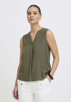 Donna che indossa una camicetta verde oliva senza maniche e pantaloni bianchi con bottoni dorati, posando con una mano in tasca contro uno sfondo semplice.