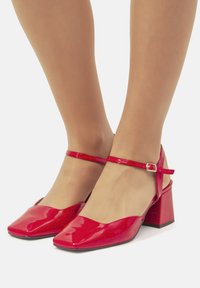 Scarpe con tacco blocco in pelle verniciata rossa, con design a punta quadrata e sottile cinturino alla caviglia dotato di fibbia metallica. Texture liscia e finitura lucida.
