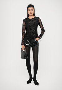 Fekete, hosszú ujjú felső áttetsző ujjakkal és flitteres virágmintával, fekete fényes rövidnadrággal és fekete harisnyával párosítva, egy kis táskával a kezében.