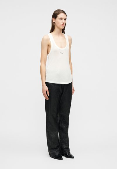 Γυναίκα που φοράει λευκό αμάνικο τοπ με στρογγυλή λαιμόκοψη και φαρδιά μαύρη υφασμάτινη παντελόνα, στέκεται με τα χέρια χαλαρά στο πλάι μπροστά σε απλό φόντο.