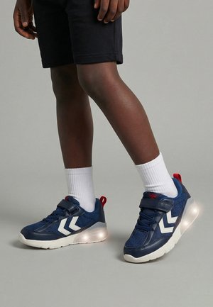 Barn iført marineblå sneakers med hvide chevronstriber, hvide sokker og sorte shorts, stående på en grå gulv.