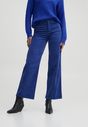 Kobieta ubrana w jaskrawo niebieskie szerokie spodnie sztruksowe, pasujący niebieski sweter z dzianiny oraz czarne czółenka na koturnie z ostrym noskiem.