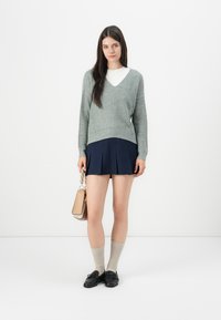 Grå strikkegenser med V-hals, kombinert med en marineblå plissert skjørt, beige knelange sokker og svarte sko, holder en liten beige veske.