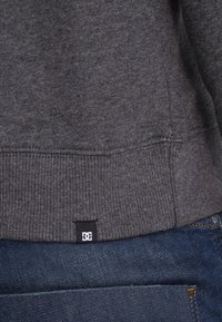 Šedá mikina s žebrovaným lemem, se malou černou značkou DC poblíž švu, v kombinaci s modrými džíny.