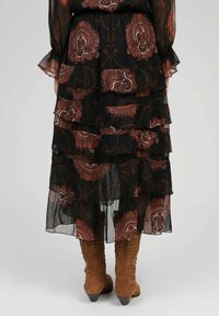 Robe maxi noire à étages avec des motifs floraux paisley dans des nuances de bordeaux et de crème. Tissu léger et transparent avec des manches longues évasées. Bottes de cowboy marron.