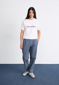 Witte t-shirt met korte mouwen met een rood en blauw New Balance-logo, gecombineerd met loslopende lichtgrijze broek en zilveren sneakers.