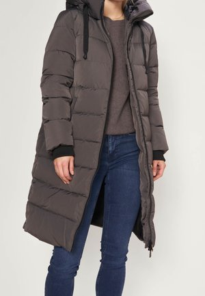 Personne portant un long manteau d'hiver matelassé gris foncé avec capuche, associé à un jean bleu et un pull marron.