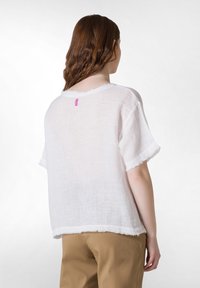 Top bianco in lino a maniche corte con orlo sfilacciato e bordi grezzi. Presenta un piccolo etichetta rosa sulla parte posteriore del collo. Indossato sopra pantaloni beige.