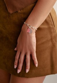 Braccialetto in catena d'argento con un ciondolo a forma di orsetto gommoso rosa e una targhetta metallica circolare. Mano appoggiata su una superficie in suede marrone.