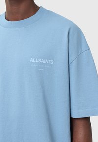 Vaaleansininen puuvillapaita, jossa on ribbikuvioitu pyöreä kaula-aukko. Edessä on valkoista tekstiä "ALLSAINTS" ja grafiikkaa. Lyhyet hihat.