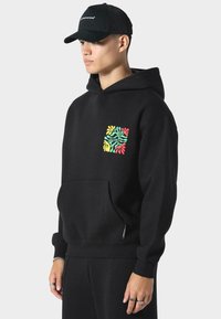 Sudadera con capucha negra con un diseño abstracto colorido en el lado izquierdo del pecho, bolsillo tipo canguro y puños acanalados. El modelo lleva una gorra negra.