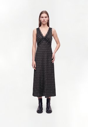 Woman wearing a black sleeveless polka dot midi dress with lace trim and black ankle boots, standing against a plain white background.