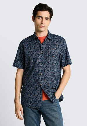 Junger Mann trägt ein blau gemustertes, kurzärmliges Hemd über einem orangen T-Shirt, mit einer Hand in der Hosentasche der Jeans.