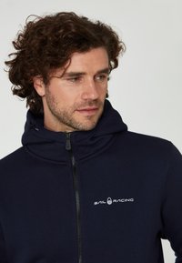 Námořnická mikina na zip z měkké tkaniny, s vysokým límcem, nastavitelnou kapucí a bílým logem "SAIL RACING" na hrudi.