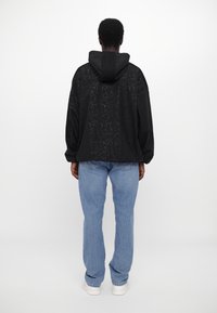 Person med en svart spräcklig hoodiejacka, ljusblå jeans och vita sneakers, som står vänd bort från kameran mot en enkel vit bakgrund.