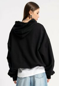 Czarny, oversize hoodie wykonany z bawełny, z dużym kapturem i opadającymi ramionami. Nosi się go na białej koszuli z luźnymi niebieskimi dżinsami.