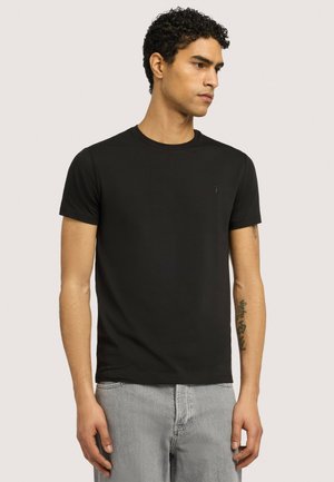 Ung mand med krøllet hår iført en ensfarvet sort kortærmet t-shirt og lysegrå jeans, stående mod en ensfarvet baggrund.