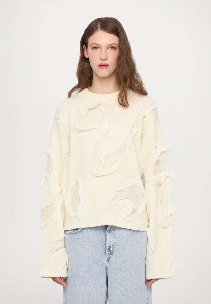 Light beige sweater with textured, frayed detailing. Features a relaxed fit and long sleeves, complementing light blue jeans.
