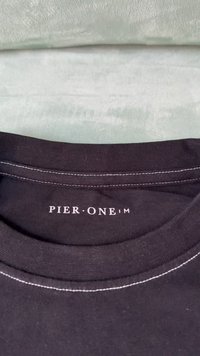 Svart bomull T-skjorte med krage og hvit søm. "PIER ONE" trykt i hvitt på den indre etiketten. Bakgrunnen har en myk, lysfarget stoff.