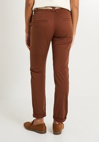 Pantalons bruns avec une coupe ajustée, dotés de poches arrière et de bas roulés. Le tissu semble lisse, agrémenté d'une ceinture tressée.