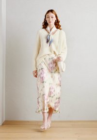 Pull en tricot crème avec un décolleté en V, superposé sur une jupe longue florale pastel. La jupe présente des teintes douces de rose et de vert, créant une silhouette fluide.