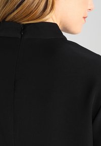 Prenda negra con un alto cuello y una cremallera oculta en la parte posterior. Textura suave y mate con líneas limpias.