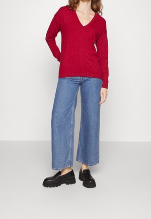 Femme portant un pull rouge à col en V, un jean bleu à jambes larges et des mocassins noirs épais, debout devant un fond blanc uni.