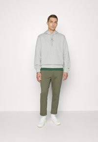 Felpa grigia con cappuccio e un accento verde in fondo, abbinata a pantaloni oliva a gamba affusolata. Modello con scarpe da ginnastica bianche.