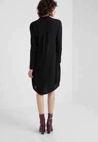Vestido negro de manga larga con cuello alto, detalle de pliegue en la parte trasera, dobladillo redondeado y combinado con botas de tobillo burdeos.