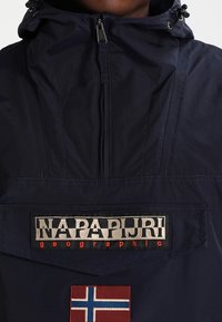 Marineblauw jack met een hoge kraag, ritssluiting en een voorzak. Heeft het "NAPAPIJRI"-logo en een Noorse vlagpatch.