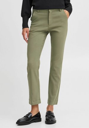 Slim-fit olive green trousers worn with black loafers, paired with a black long-sleeve top, hands in pockets, standing on a plain background.