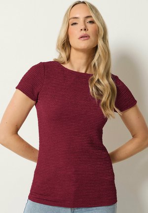 Blondynka z luźnymi falami, ubrana w bordową teksturowaną bluzkę z krótkim rękawem i jasnoniebieskie dżinsy, stojąca na jednolitym tle.