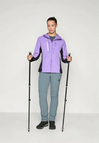 Chaqueta de senderismo morada y negra con cremallera y logo, combinada con pantalones gris claro y botas de senderismo negras. Sostenía bastones de trekking.