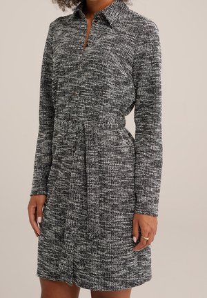 Vrouw draagt een lange mouwen, zwart-wit gestructureerde blousejurk met riem, knopen en kraag, staand tegen een neutrale achtergrond.