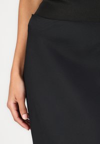 Jupe noire avec une taille ajustée, dotée d'un tissu lisse et texturé et d'une silhouette près du corps, main reposant sur le côté.