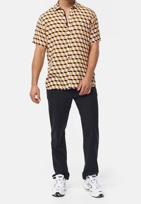 Chemise à manches courtes avec un motif géométrique en marron, crème et noir ; associée à un pantalon sombre et des baskets blanches.
