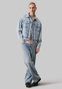 Giubbotto di jeans azzurro chiaro con due tasche frontali, abbinato a una maglietta grigia chiara e jeans azzurri baggy. Scarpe nere chunky.