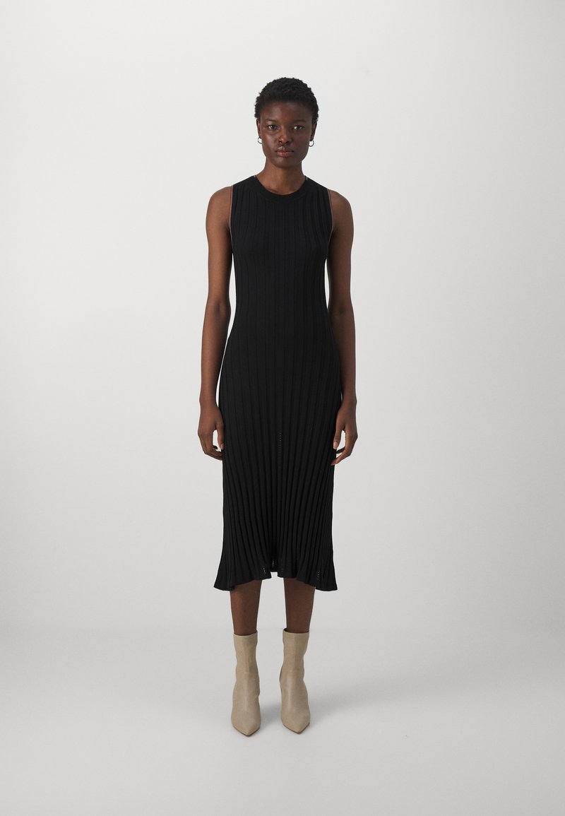 Robe midi noire à côtes avec un design sans manches, présentant une silhouette ajustée et un ourlet évasé. Portée avec des bottines beige à la cheville.