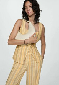 Beige leather handbag with a curved shape, carried by a person wearing a yellow-striped suit, featuring wide-leg trousers and a fitted vest.