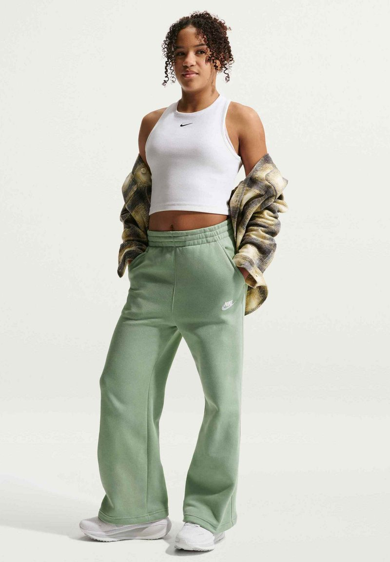 Jeune femme portant un crop top blanc Nike, un pantalon de survêtement vert Nike, des baskets blanches et une veste à carreaux drapée sur ses bras.