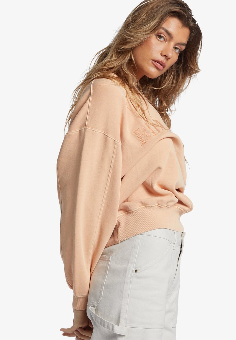 Sudadera de gran tamaño en color durazno con texto bordado, con hombros caídos y puños acanalados, combinada con pantalones de tiro alto en gris claro.