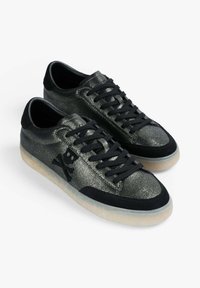 Par de zapatillas negras de corte bajo con acabado metálico, cordones negros, logo de calavera y huesos cruzados, y suelas de goma translúcidas.