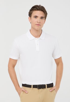 Maglietta polo bianca in cotone con colletto e apertura a due bottoni, maniche corte, abbinata a pantaloni beige e una cintura intrecciata nera.