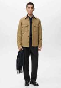 Tan wool blend shirt jacket with two front pockets, worn over a black polo shirt, paired with black trousers and a black scarf. Black shoes.