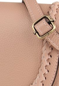 Details van lichtroze leren handtas met een gevlochten riem en messing beslag met contrasterende stiksels. Gestructureerde afwerking, zachte uitstraling.