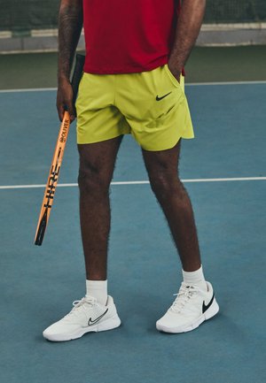 Nike tennis shorts in neon yellow with side pockets, paired with white Nike shoes and black accents. Holding an orange tennis racket.