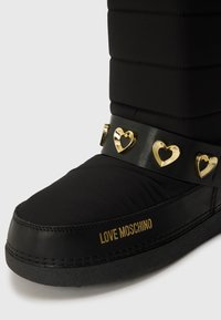Zwarte winterlaarzen met een gequilte nylon bovenkant, een dikke rubberen zool en een decoratieve zwarte band met gouden hartaccenten. Gouden logo-detail.