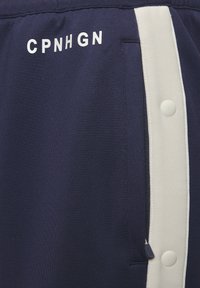 Pantalones cortos deportivos en color marino con franjas laterales blancas, que cuentan con un bolsillo con cremallera y el texto "CPNHGN" impreso en blanco en la parte delantera.