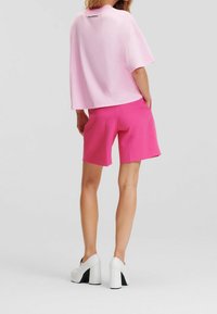 Žena oblečená v světlé růžové tričko s krátkým rukávem, jasně růžových šortkách a bílých platformových lodičkách, stojí s rukama v kapsách.