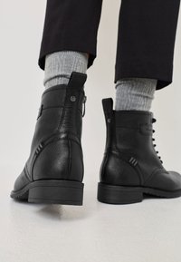 Isik, kellel on jalas mustad nahkjalatsid külgkangide ja hallide sokkidega, seisab valgel tekstuurilisel põrandal.